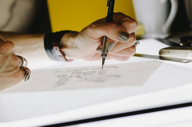 Close-up photograph of a person sketching a design on paper with a mechanical pencil, with a yellow notebook, white mug, and smartphone on the desk.